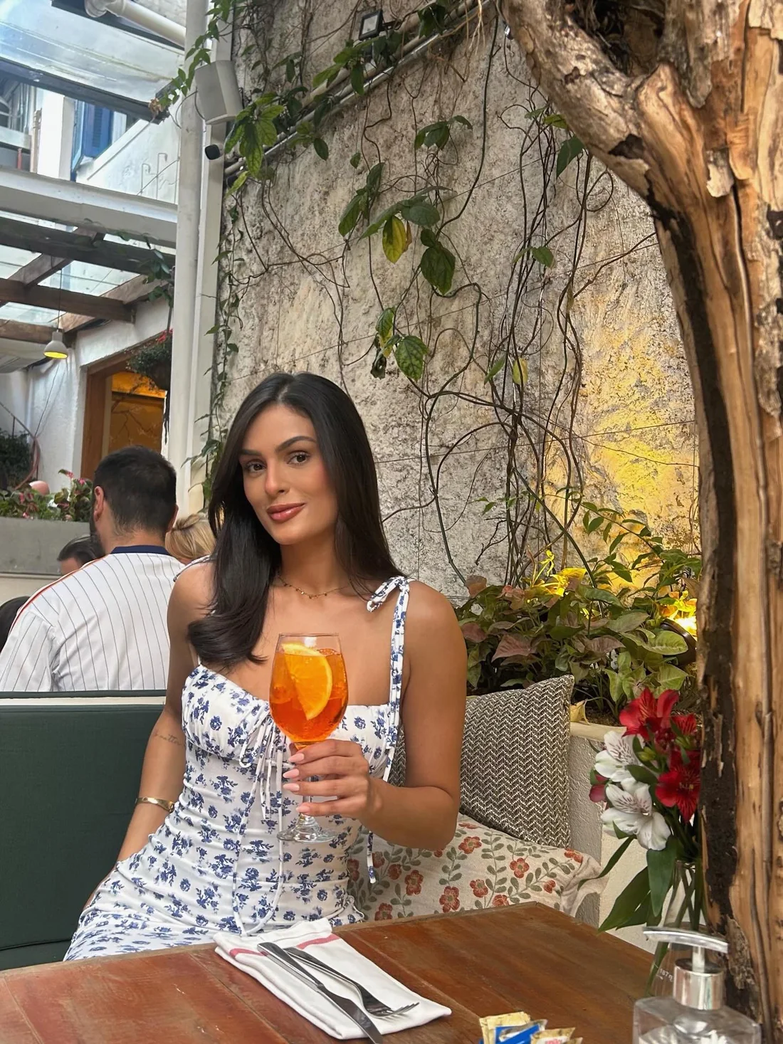 Débora Xavier de vestido floral, segurando um drink laranja em um jantar em restaurante com jardim