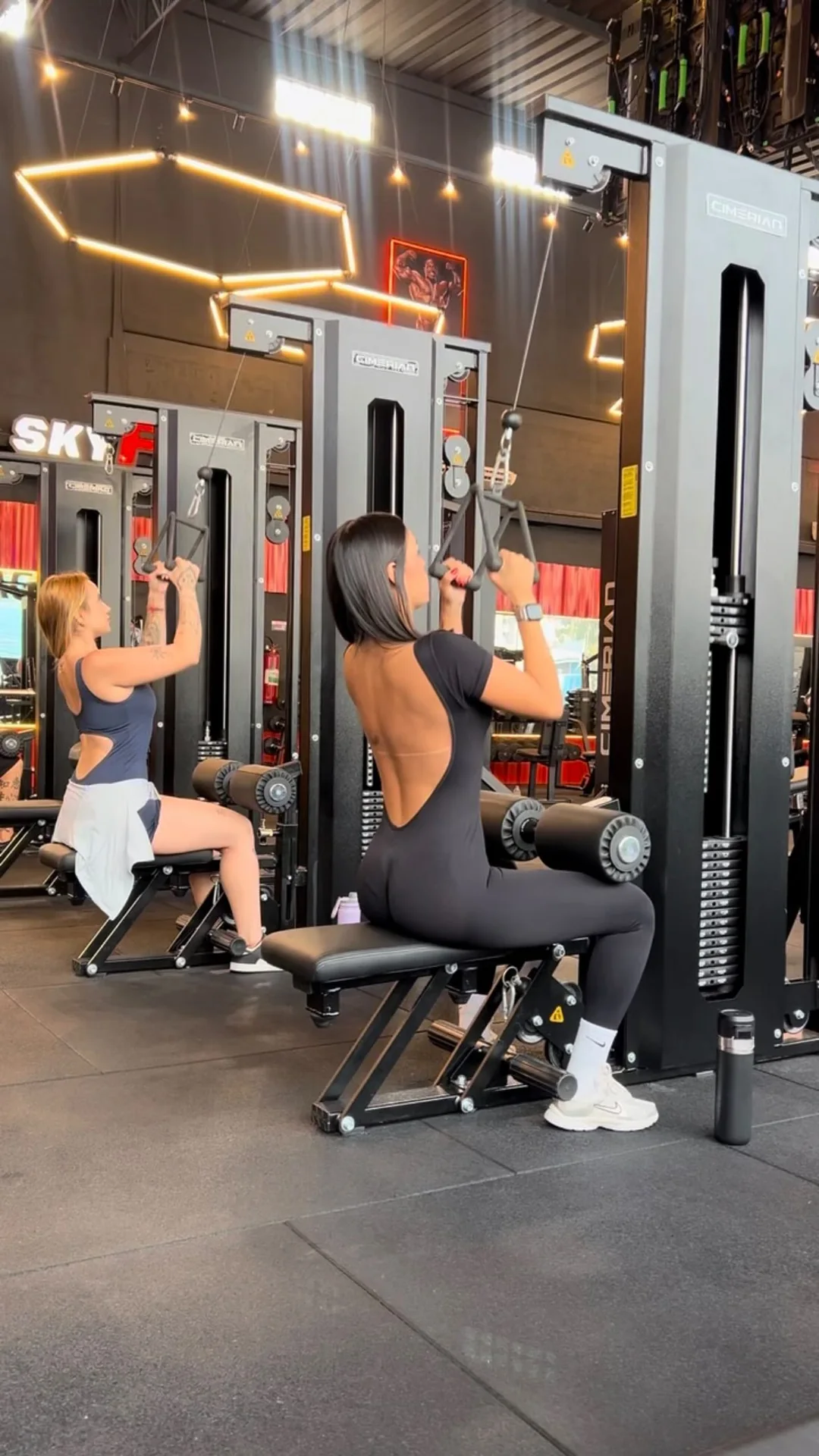 Débora Xavier de macacão preto com as costas à mostra, treinando na polia da academia