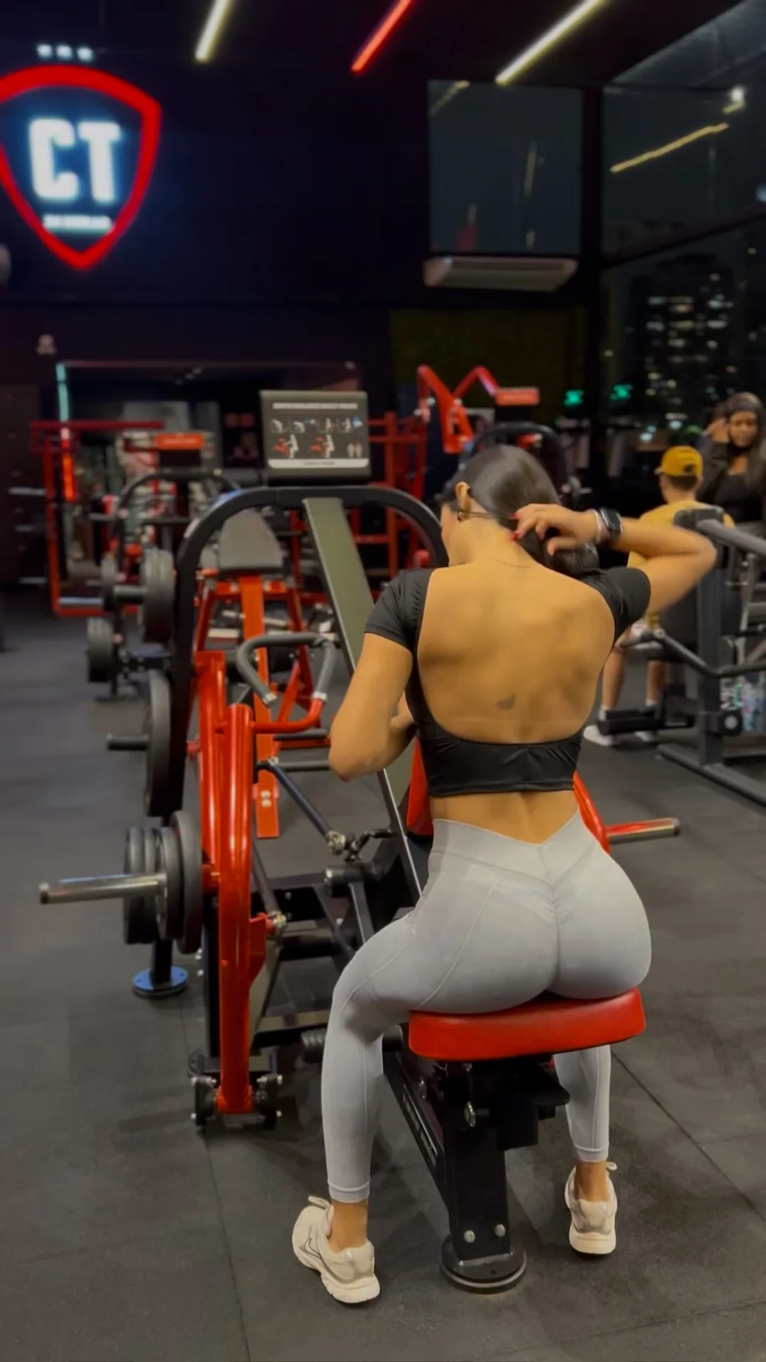 Débora Xavier de costas, de legging cinza, na máquina de puxada da academia