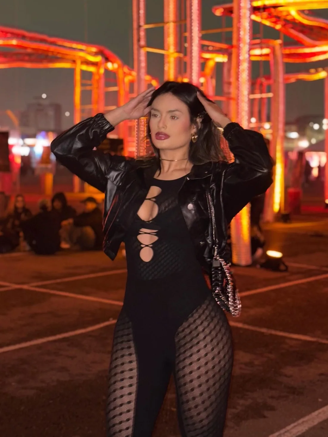 Débora Xavier de jaqueta de couro e macacão preto rendado, com a mão no cabelo, entre estruturas alaranjadas de festival